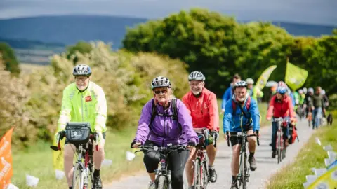 Steve Barber People cycling along a narrow path of bay Cycle Way on its launch day in 2015. It is surrounded by grassland and trees, with a hill in the distance.