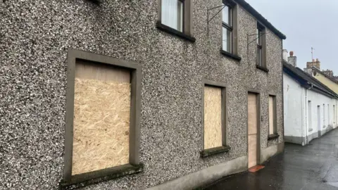 House in Broughshane showing wooden boarded up windows at the bottom. The house has a pebble dash effect.