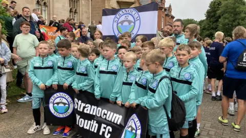 Shows boys in green football strips and carrying a banner of Stranocum Youth FC