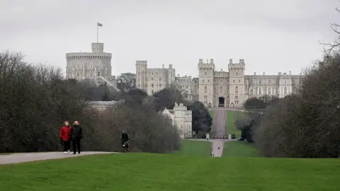 Getty Images Windsor Castle seen from the Long Walk in Windsor Great Park. A couple of people can be seen walking together on the path in coats. Another person on a bike is riding a short distance behind them. The path is flanked by well-tended grass and rows of trees.