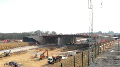 The structure of a large bridge on a construction site, sitting above orangey churned up earth on a clear day. There are trees to the left of the bridge and diggers and other construction vehicles can be seen in front of it.