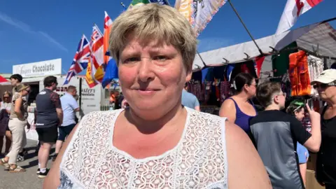 A woman with short light hair wearing a white and blue top looks at the camera on a sunny day.  She is standing in front of a food stall.  There are crowds of people in the background and row of flags which including the Union flag. 