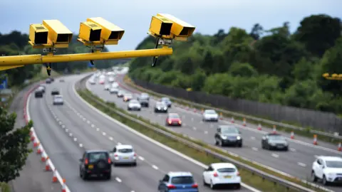 PA Yellow ANPR cameras in the foreground of the photo, below them is a motorway with cars on it, with trees in the background