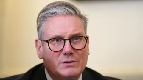 PA Media Sir Keir Starmer is pictured sitting in a room facing away from the camera. He is wearing black glasses and a black suit and a white shirt underneath