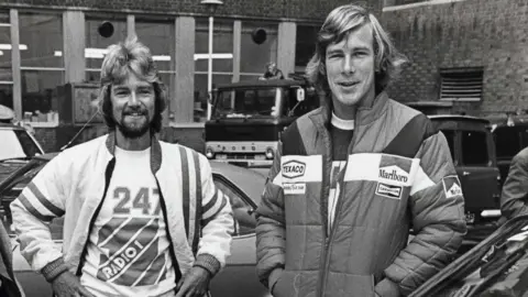 John Curtis/Shutterstock A black-and-white mid-shot of two men in their late twenties, standing side-by-side facing the camera, pictured from the waist up. The man on the right is taller by about six inches, although also standing slightly closer to the camera. Both men have long, fair, wavy hair, although the man on the left is bearded while the man on the right is clean-shaven. The man on the left wears a white jacket with dark stripes, with an open zip-up front showing a white t-shirt beneath with a large Radio 1 logo printed on it. The man on the right wears a jacket which shows up as a mid-range grey in the photo, bearing logos of motor racing sponsors such as Texaco and Marlboro.