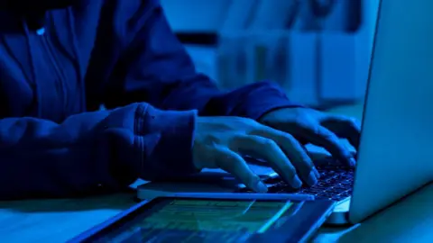 A blue-lit room with a person in a hoodie typing on a laptop