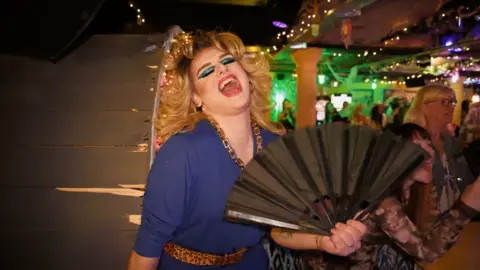 A drag queen with blonde, Farrah Fawcett-style hair laughs at the camera as she fans herself with a black fan. She wears a blue dress with a leopard print belt