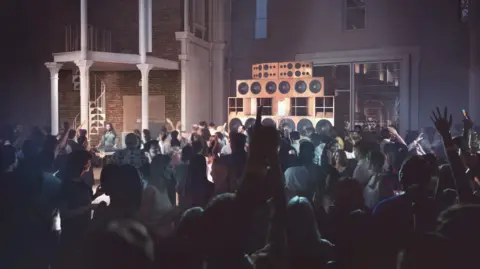 Secchi Smith Asif Khan A large crowd dances in a dimly lit indoor venue with tall speakers stacked near a brick wall, as the spiral staircase in the background overlooks the lively market atmosphere.