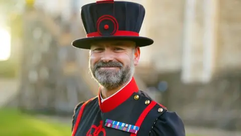 Ryan Brown with a short cropped beard stands smiling at the front of the tower of London. He is wearing a ceremonial blue and red tunic and a tall blue hat. The hat is trimmed with red. A Good Conduct medal for long service is pinned on the chest of his tunic.
