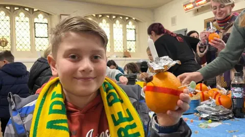 Andrew Turner/BBC Jonah holds one of many Christingles he made during a workshop at Great Yarmouth Minster on Monday. Jonah, has blonde hair and is wearing a yellow and green Norwich City Football Club scarf. 