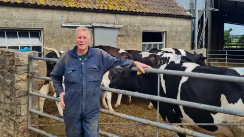 A farmer in blue overalls leans against a metal gate, with a herd of black and white cows gathered behind him. The backdrop features an old stone barn with a moss-covered tiled roof. Another farm building is visible to the right under a partly cloudy sky.