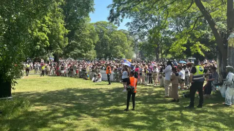 Lily-May Symonds/BBC Crowds of people in a park during a carnival celebration. The crowds are standing behind a fence looking toward a stage. 