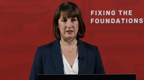 Getty Images Rachel Reeves stands in front of a red wall at a press conference