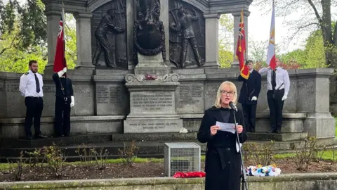 Sharon Coveney speaking in front of the Titanic Engineers' Memorial in East Park the memorial has an angel stood tall in the centre and she is flanked by workers.