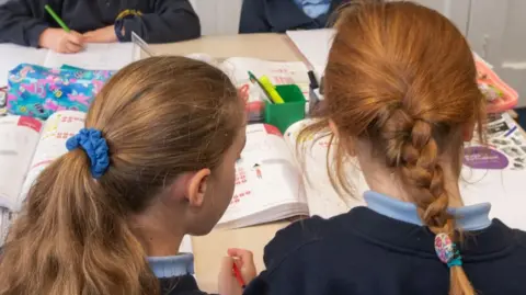 A desk covered in open school text books on top of a table at which four pupils are seated. The foreground shows the backs of the heads of two girls with their hair pulled back in pony tails. In the background two pupils sit facing the camera with only their hands and parts of their trunks showing. All four students are wearing dark blue sweatshirts with light blue collared tops underneath.