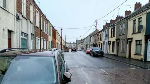 A street with terraced houses on both sides. Some cars are parked. It's been raining.