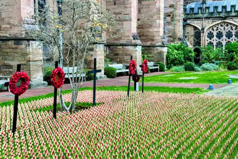 BBC Weather Watchers / Just Smile A poppy display outside Hereford Cathedral. Hundreds of small wooden crosses decorated with a small poppy are standing in a patch of grass, with four wreaths on larger black crosses behind them. The cathedral is in the background