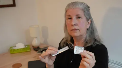 A middle-aged woman with long grey hair holds up two silver-coloured blister packs of round white tablets as she sits in a dining room with white walls. She is wearing a black top and has a serious expression on her face.