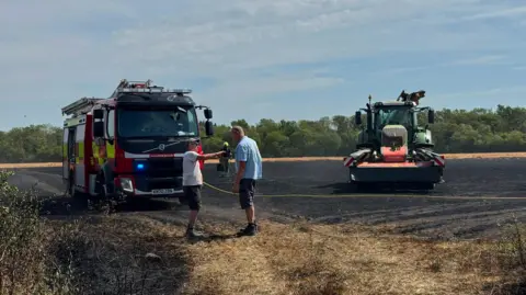 Thames Valley Police Mounted Section A fire engine and a tractor in a field blackened by a fire. Two men are talking to each other in the foreground. There is a line of trees in the background.