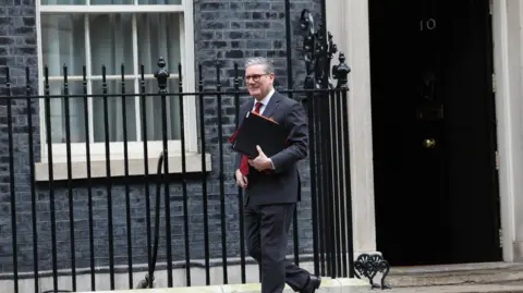 EPA Prime Minister Sir Keir Starmer leaves 10 Downing Street. The black door with 10 on it can be seen in the background as Sir Keir walks away. He is wearing a dark suit and glasses and is holding a binder. 