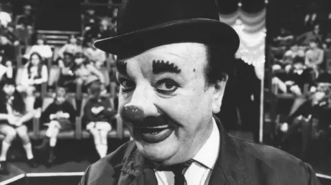 Black and white image of Charlie Cairoli in front of a crowd of children in a theatre. He is wearing a bowler hat, suit and has a big clown nose, large eyebrows, a moustache and dark lips