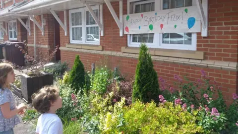 Julie Ricau A young boy and girl stand in front of a front garden and look at a colourful sign that says "happy birthday Felix"