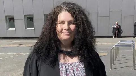 Diane Barnes A middle-aged white woman standing outside a large grey building. She is wearing a graduation gown and has black hair. She is smiling at the camera.