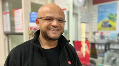 Sid Patel is smiling as he stand in front of the post office counter. He is wearing rimless glasses and has a beard. He is wearing a post office black jacket. 