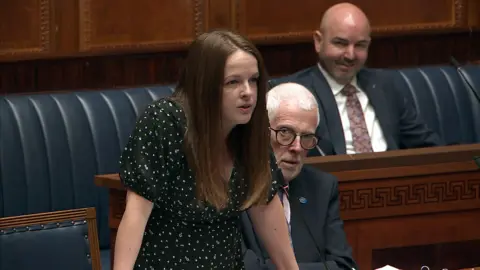 NI Assembly Nuala McAllister, she has long brown hair and is wearing a black floral top, stood in the NI Assembly chamber, there are blue leather seats and wooden tables