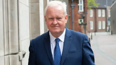 Staffordshire County Council Councillor Ian Parry is wearing a white shirt and blue suit, with a blue tie that has white spots on it. He is stood in a street, with red brick buildings in the background.