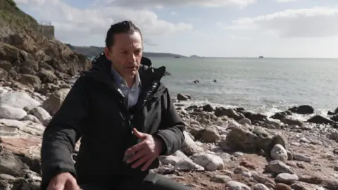 Dr Gavin Tilstone is in a black coat, black trousers and white shirt. He is sat on a rock on a rocky beach. He is facing the camera. The sea is in the background. The sky is blue with some white clouds. 