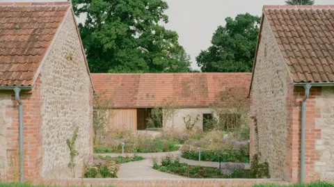 Lorenzo Zandri Un ensemble de bâtiments en pierre d'un seul étage autour d'un jardin avec des allées sinueuses et des parterres plantés de graminées ornementales et de fleurs aux teintes roses et violettes.