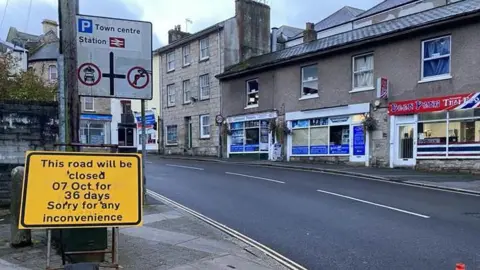 A road in Redruth. At the front of the picture there are two signs. One is a road sign with directions to the town centre and the other says 'This road will be closed on 7 October for 36 days. Sorry for any inconvenience'. There is a row of shops on the opposite side of the road. 