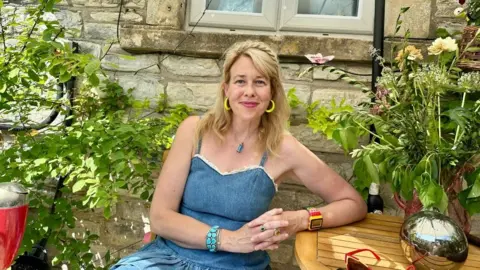 A woman is sitting in her garden next to a wooden table. There are plants around her. She is wearing a blue top and yellow earrings.