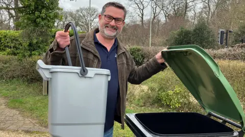 Jon Neal is standing on a gravel drive with trees and bushes in the background. He is holding a small grey caddy in one hand while standing next to a black and green wheelie bin, which he is holding open. He is wearing blue jeans, a blue jumper and a brown jacket. He is wearing glasses.