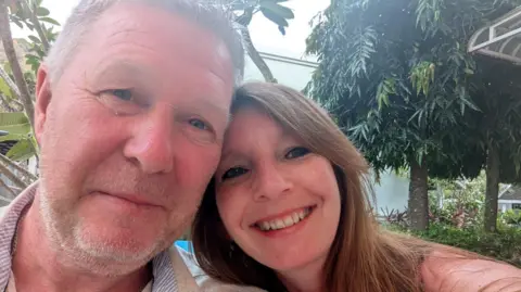 Supplied A man and woman smiling in a selfie, their heads touching. He is on the left with grey hair and stubble, she is to his right and has brown long hair. Behind them are tropical-looking trees. 