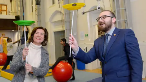 Arts Council of Wales First Minister Eluned Morgan and Culture Minister Jack Sargeant spinning plates during a circus workshop. Eluned Morgan is holding a green plate on a stick while Jack Sargeant is holding a yellow plate.