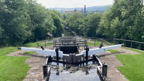 Heidi Tomlinson/BBC A picturesque section of a canal, with walkways bridging over the water. The canal is flanked by grass verges and trees on both sides.