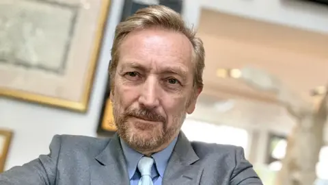 Michael Van Clarke Close up of a man with strawberry blond short hair, beard and moustache, wearing a light grey suit, blue shirt and paler blue tie. He has a neutral expression and you can see a picture frame blurred in the background.
