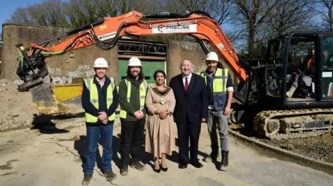 Crawley Borough Council Three people in high-vis vests and a man and a woman in smart clothing at a building site with a small digger behind them