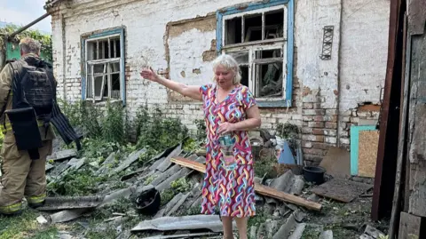 A woman with white hair in a pink dress stands looking destressed with her arm out infront of a bombed out building with a Ukrainian soldier to her left 