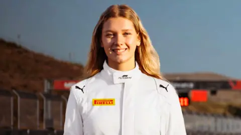 Rachel Robertson wearing a white racing suit with various sponsors logos on it. The metal fencing of a racetrack can be seen in the background.