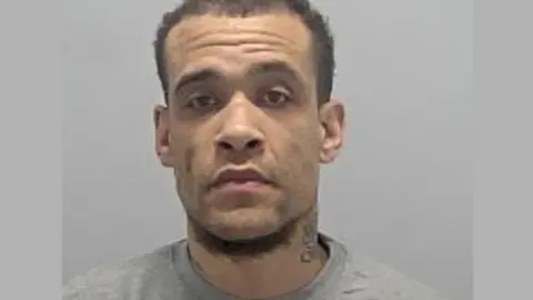 A police mug shot of Callun Bermingham (tbc sp). He has short black hair and a light beard. He is wearing a grey sweatshirt.