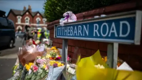PA Media Bunches of flowers and teddies lined up against a wall next to a road sign reading Tithebarn Road in Southport.