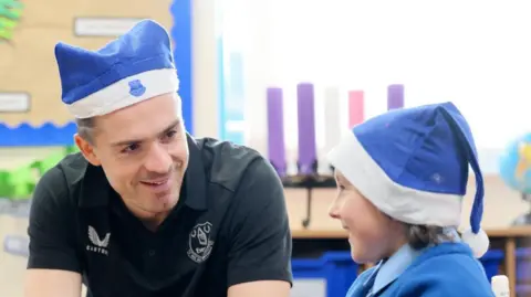EFC Jack Grealish, who wears an Everton blue santa hat and black t-shirt, smiles as he chats with Georgie, who wears the same hat and blue school sweater. They are sitting at a table in a classroom with colourful items pinned up on the wall behind.