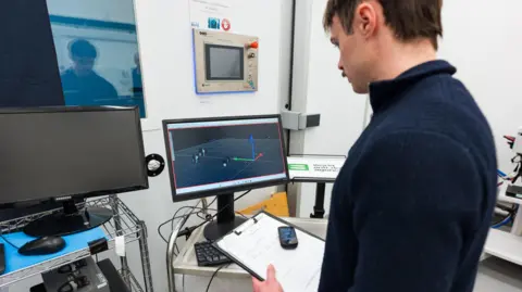UNIVERSITY OF KENT A researcher wearing a blue jumper looks at a clipboard and several computer screens in a laboratory.