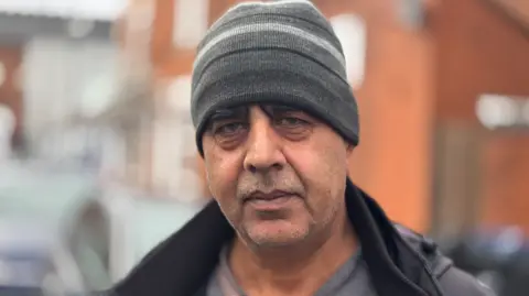 Mr Rehman is wearing a grey hat and black coat and is stood in the street