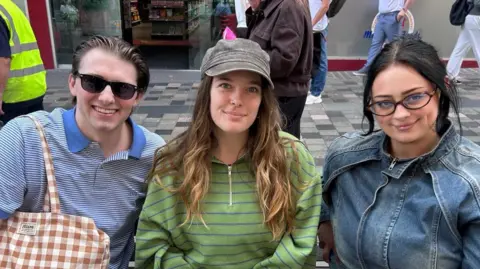 A man and two woman on a bench in Glasgow. The man is wearing sunglasses and a striped top. The woman in the middle is wearing a green striped fleece and hat and the woman on the right is wearing a denim jacket and glasses.
