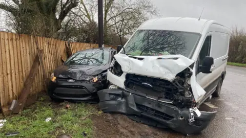A heavily damaged white van and black Ford car rest beside a roadside fence after a collision, with debris scattered across the muddy verge and road.