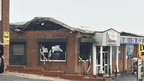 A red brick commercial building pictured from across the street in the daytime. The building is blackened in places from fire damage and there is no glass in the windows.
